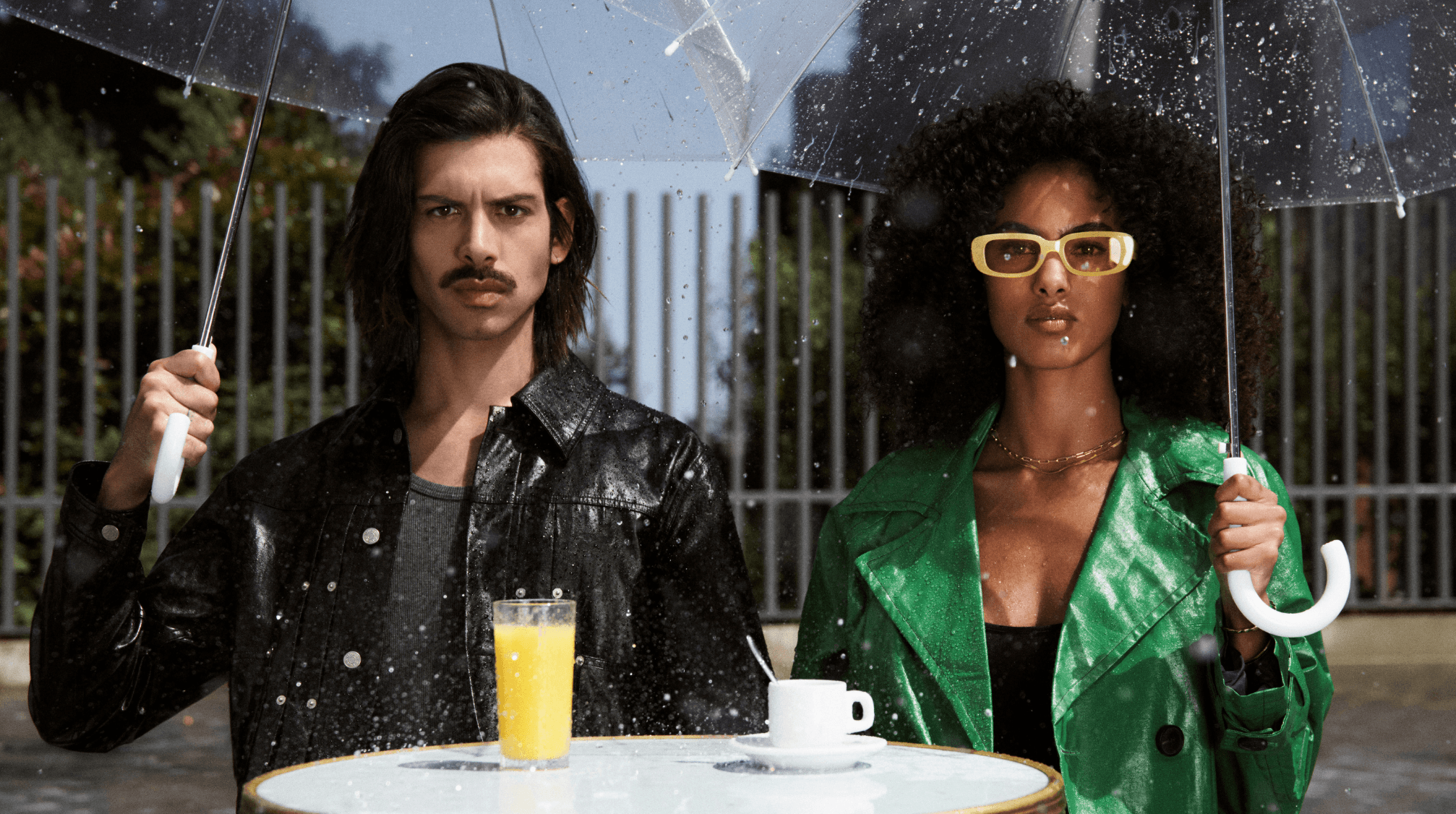 homme avec une coupe cheveux long et une femme avec les cheveux très bouclés sous des parapluies sous la pluie à une terrasse de café parisienne