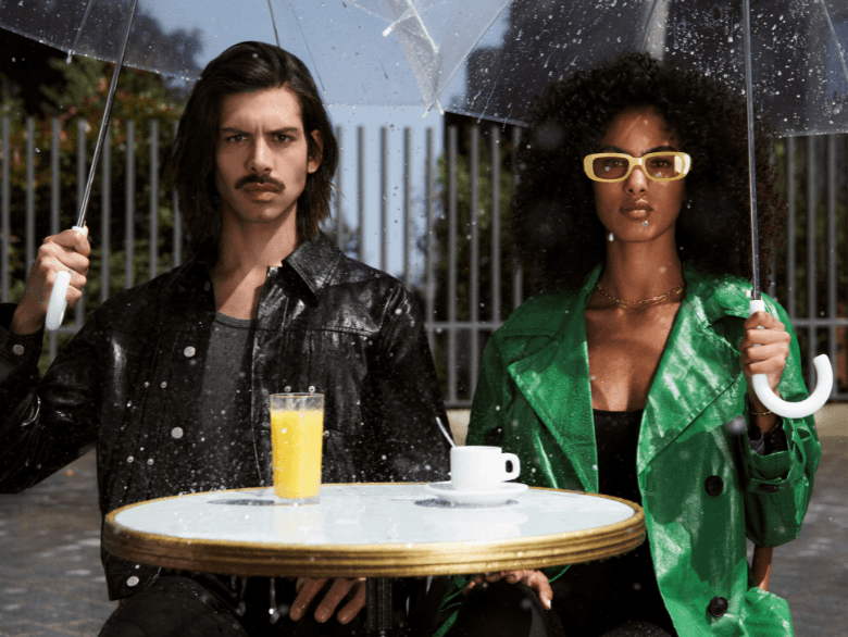 homme avec une coupe cheveux long et une femme avec les cheveux très bouclés sous des parapluies sous la pluie à une terrasse de café parisienne