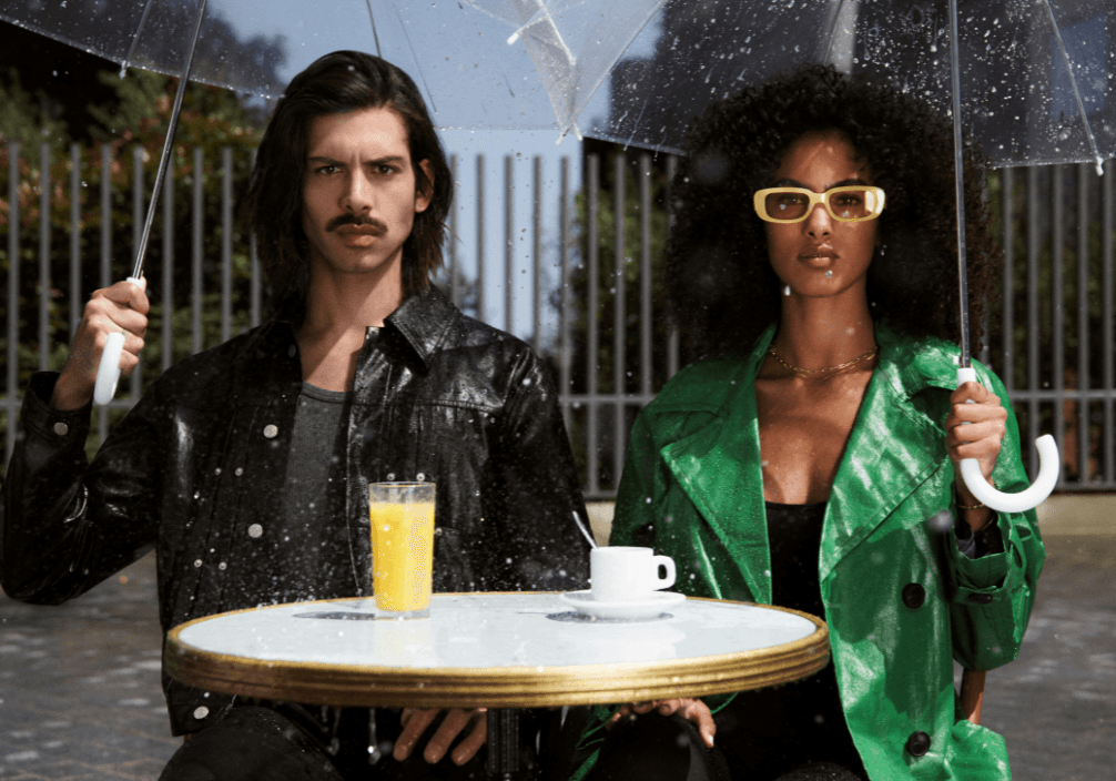 homme avec une coupe cheveux long et une femme avec les cheveux très bouclés sous des parapluies sous la pluie à une terrasse de café parisienne