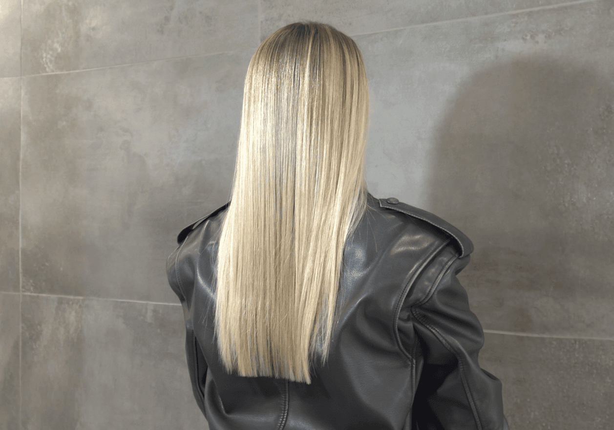 Vue de dos d'une personne aux longs cheveux blonds parfaitement lisses et droits, tombant de manière uniforme sur le haut du dos. La chevelure blonde claire et lumineuse présente une texture soyeuse. La personne porte une veste en cuir noir structurée et se tient devant un mur en carrelage gris foncé.