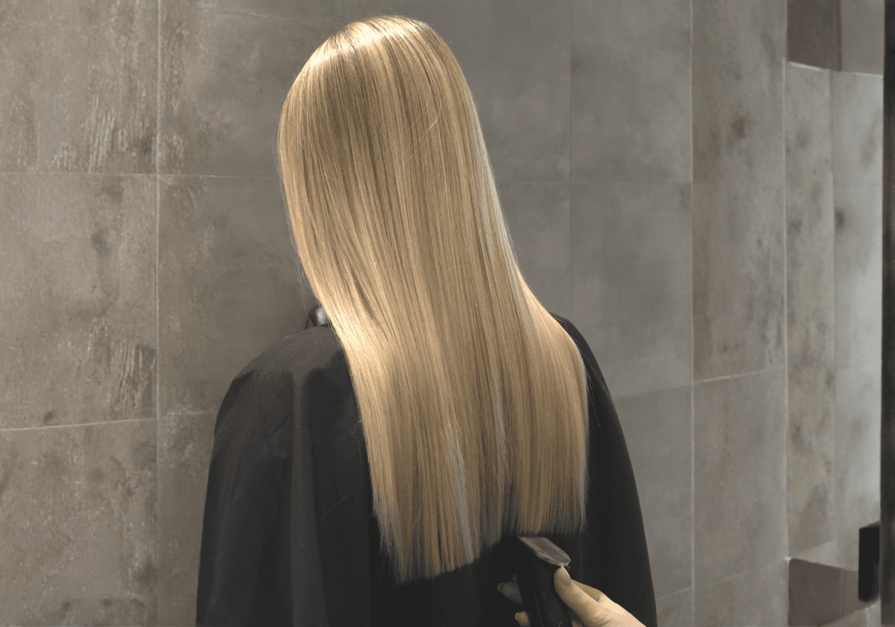 Vue de dos d'une personne aux longs cheveux blonds parfaitement lisses et droits, tombant de manière uniforme sur le haut du dos. La chevelure blonde claire et lumineuse présente une texture soyeuse. La personne porte une veste en cuir noir structurée et se tient devant un mur en carrelage gris foncé.