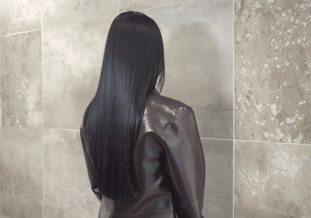 Soin Botox capillaire Jean Louis David sur cheveux longs et lisses, effet brillance intense et fibre capillaire réparée. Vue de dos d'une personne aux longs cheveux noirs parfaitement lisses et soyeux, tombant en pointe au milieu du dos. La chevelure présente une brillance intense et uniforme sous la lumière. Elle porte une veste en cuir marron et se tient devant un mur en carrelage beige.